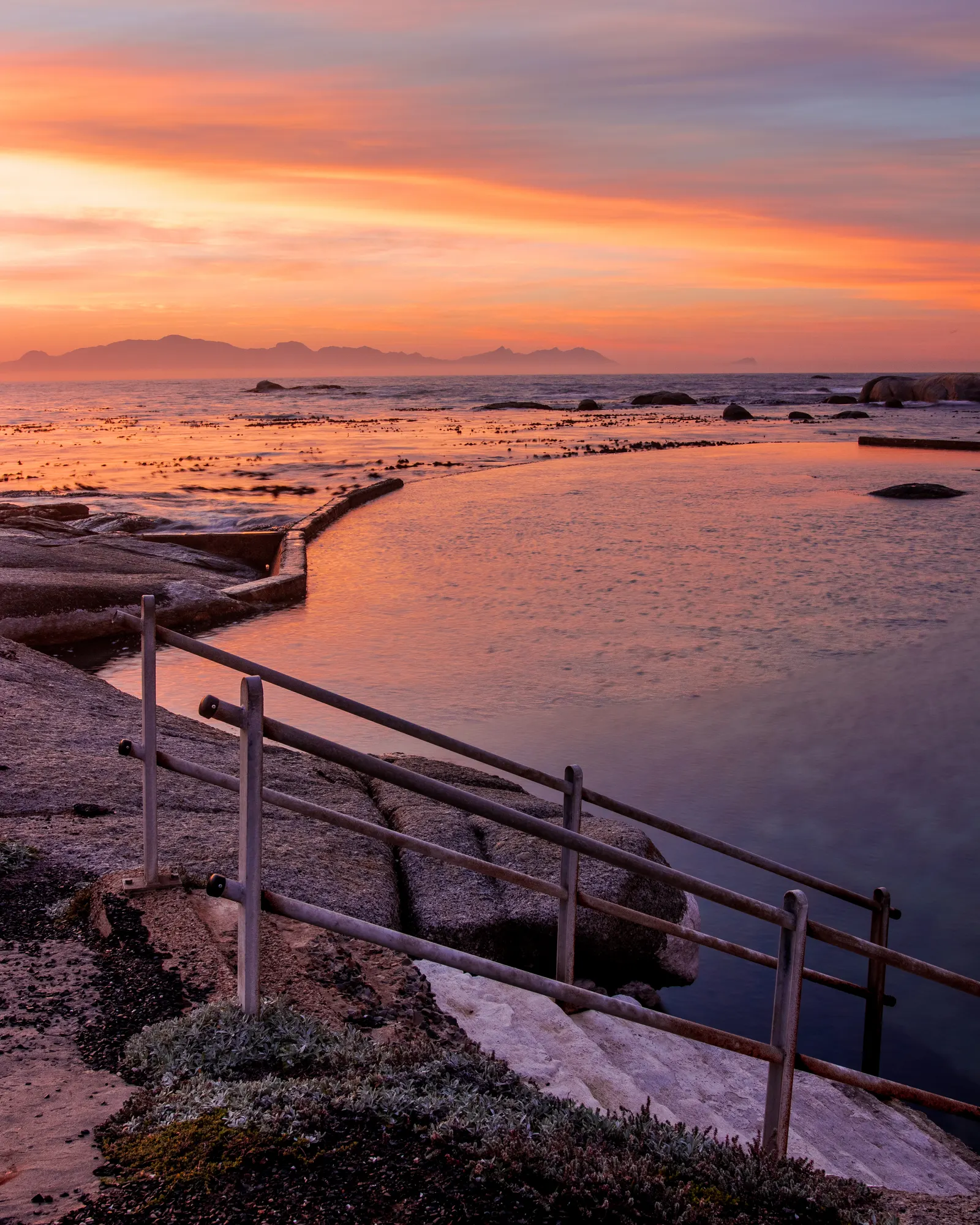 Miller's Point tidal pool at sunrise, iron railings leading toward an orange and gold sky with the Hottentots Holland mountains on the horizon