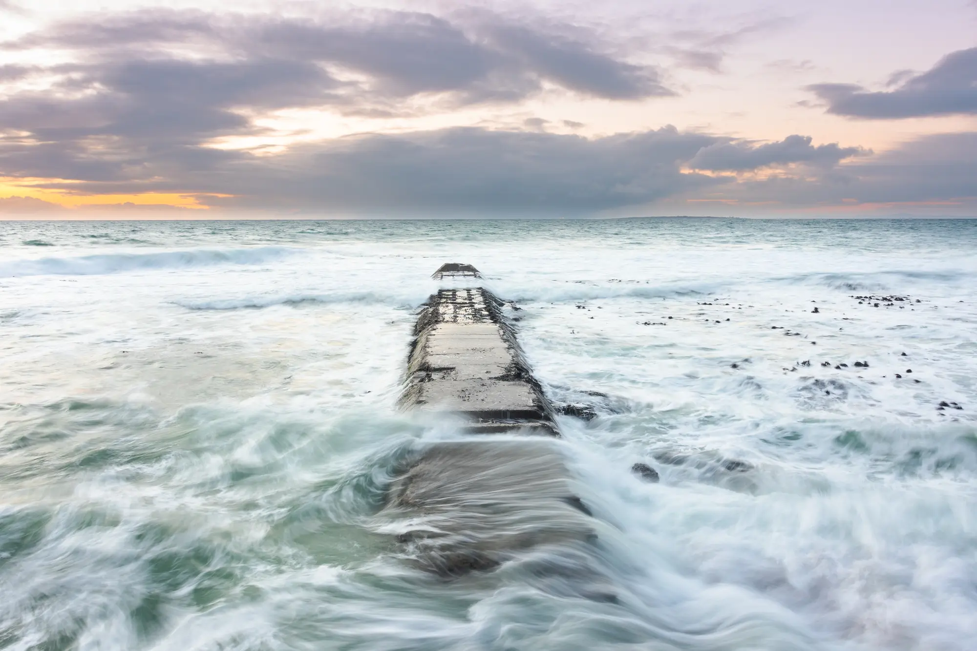 A crumbling jetty at Mouille Point reaching into a churning sea at sunrise, waves breaking over it from both sides