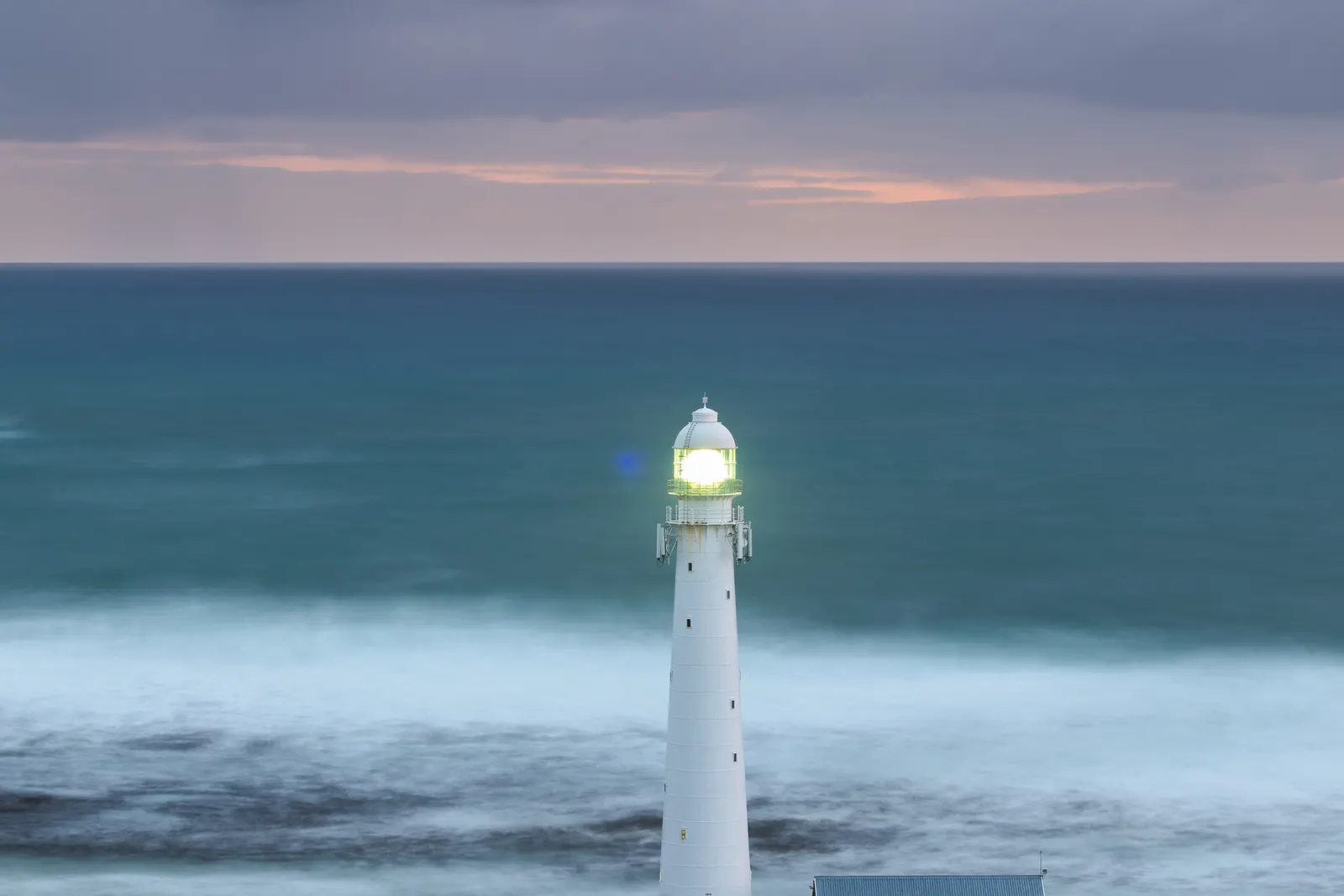 Slangkop Lighthouse with its beam lit as a long-exposure storm rolls in from the Atlantic, waves reduced to mist around its base