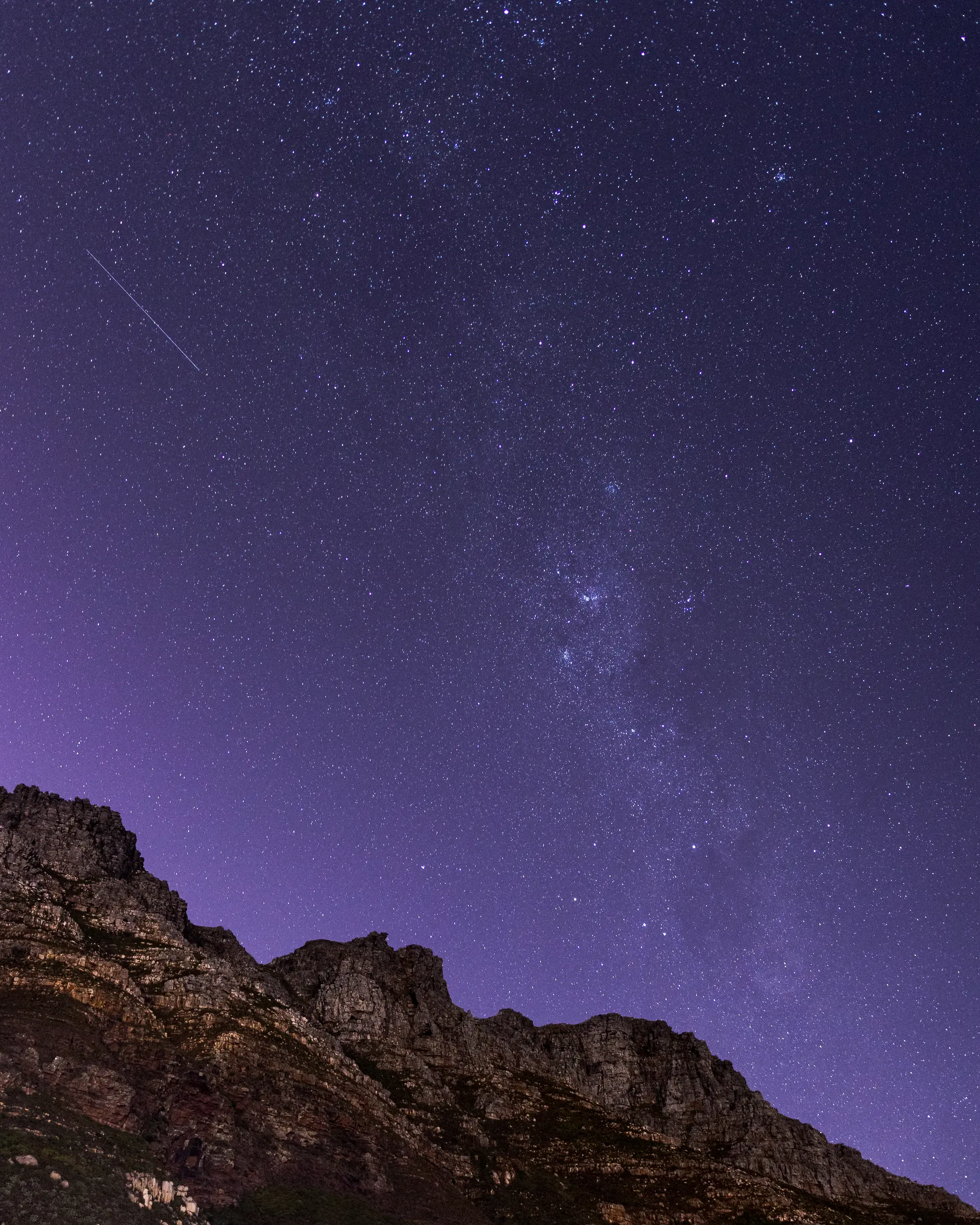 The Milky Way rising above the jagged ridgeline of the Twelve Apostles, a satellite trail crossing the deep violet sky