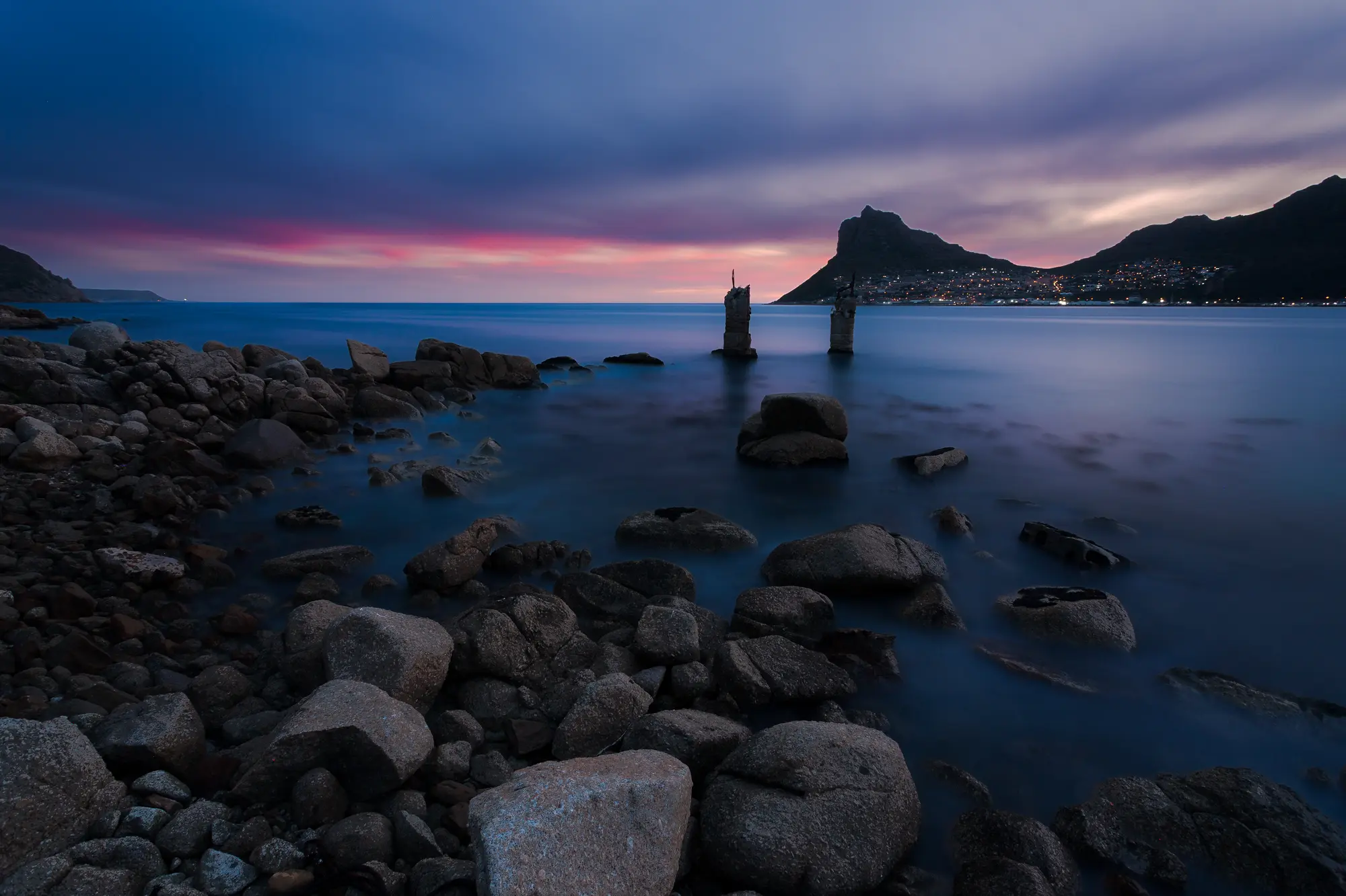 The rusted remains of Hout Bay's old iron ore jetty rising from still evening water, the Sentinel mountain sharp against a pink and violet sky